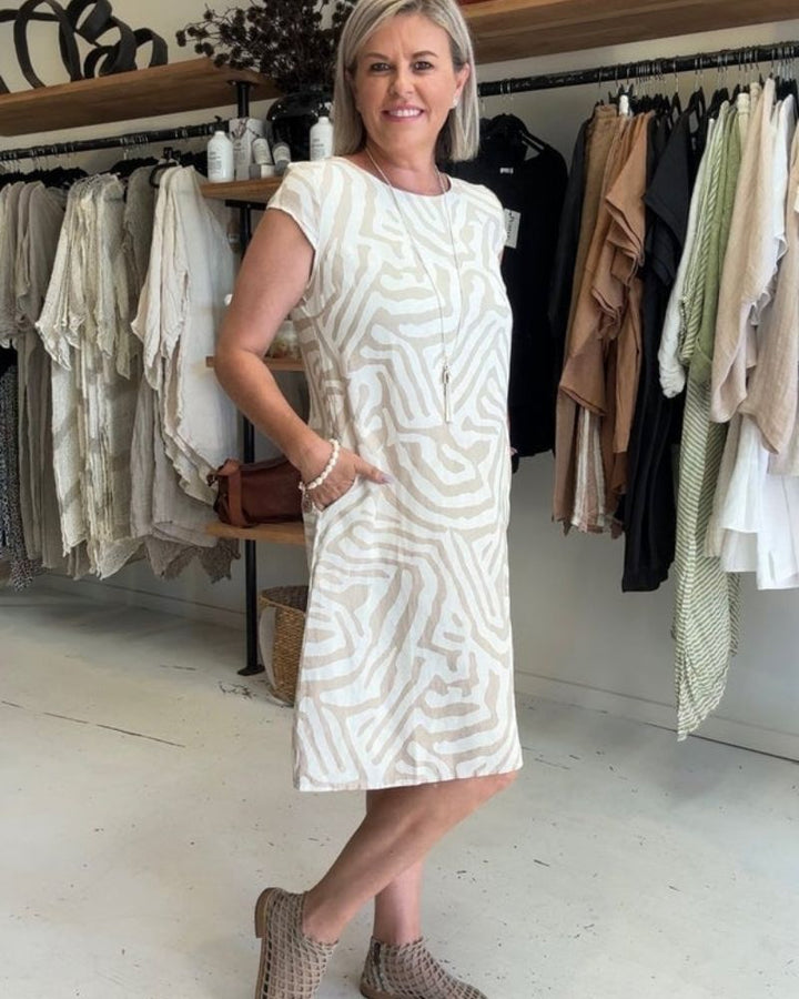 Vrouw in een witte zomerjurk met abstracte print poseert casual in een modewinkel met kledingrekken.