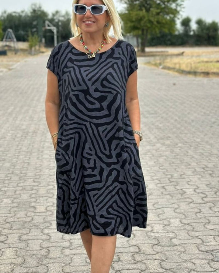 Blonde vrouw draagt een zwarte zomerjurk met abstracte print en ketting, wandelend op een buitenpad.