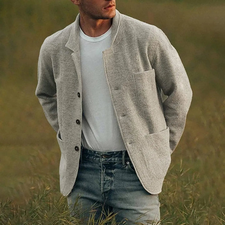 Man in een lichtgrijze casual blazer, gecombineerd met een wit T-shirt en jeans, staand in een groen veld