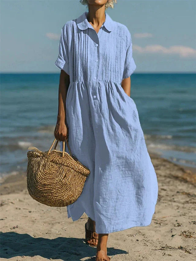 Model toont een lichtblauw losvallend hemdblusenkleed met knoopsluiting aan de kust.