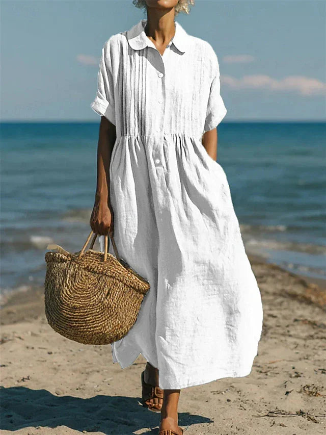 Vrouw draagt een wit hemdblusenkleed in midi-lengte met korte mouwen op het strand, met een rieten tas.