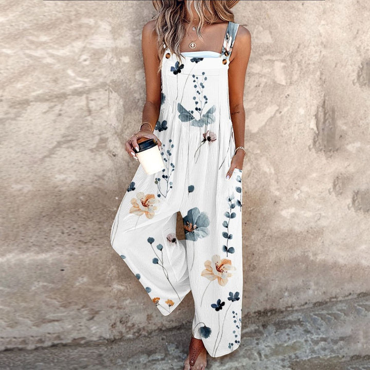 Vrouwelijke model poseert met wit zomer-jumpsuit met bloemenprint tegen een rustieke achtergrond.